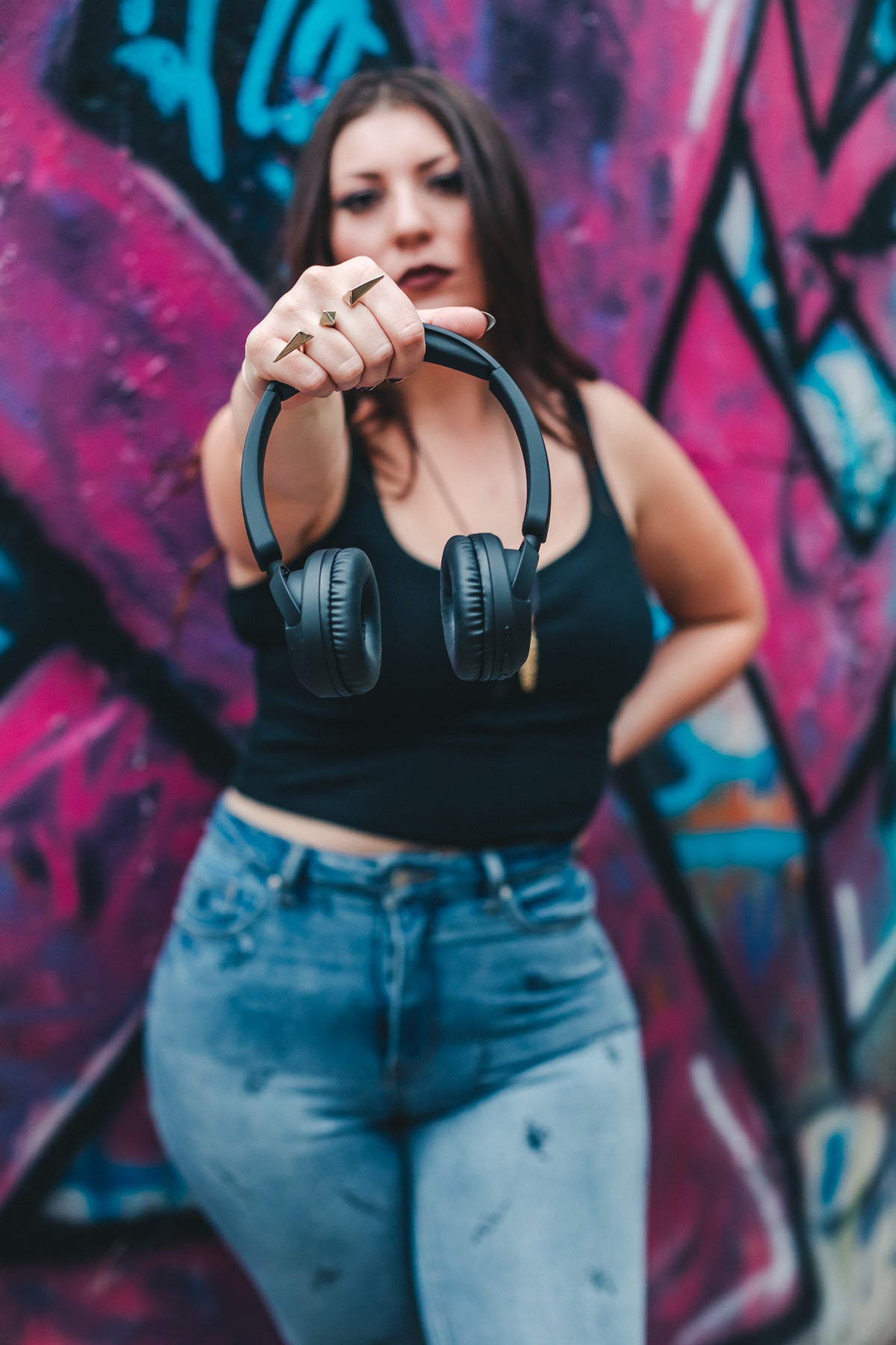 Woman standing in front of graffiti wall holds headphones out towards camera